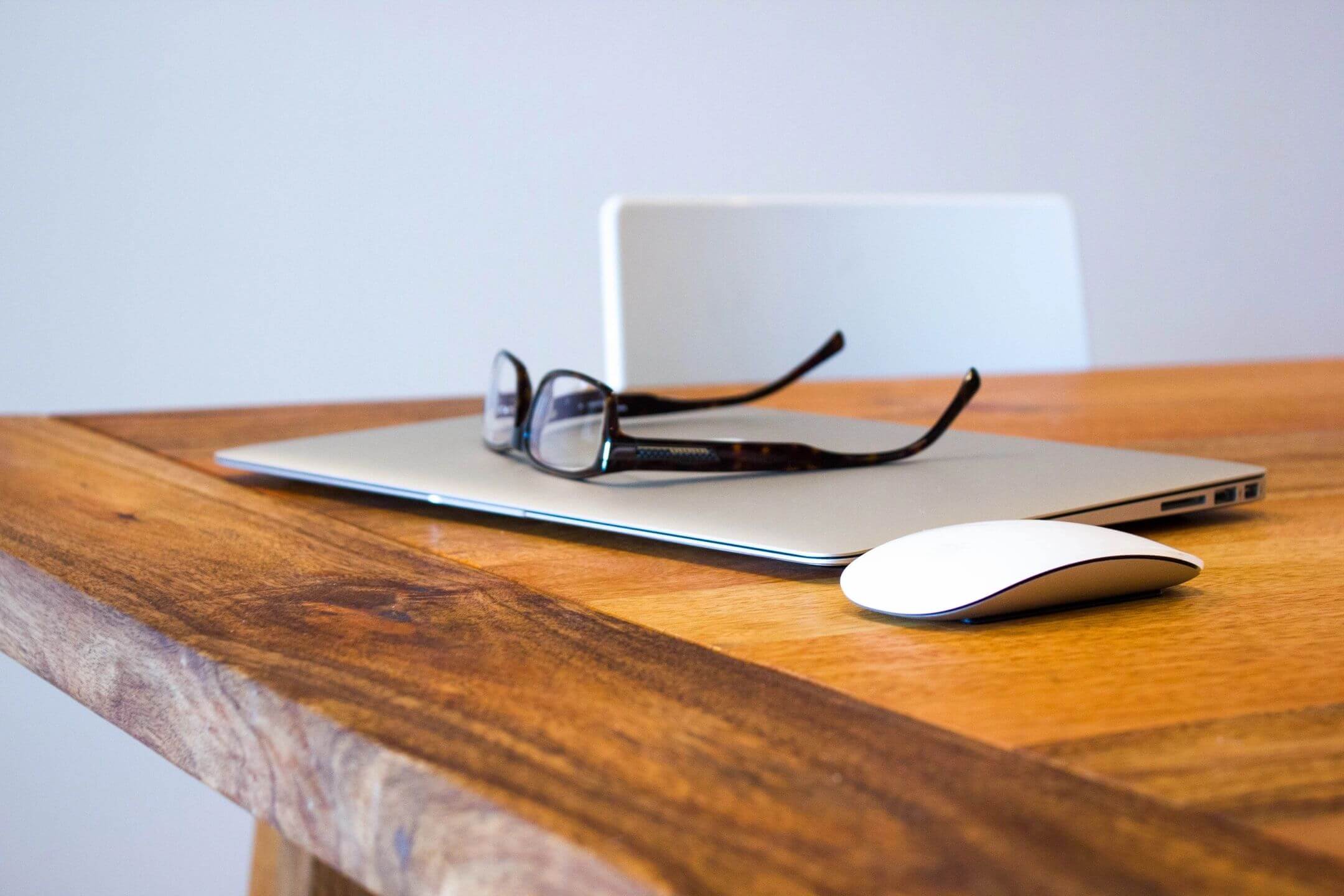 Glasses resting on a closed laptop on a wooden table with a mouse beside it.