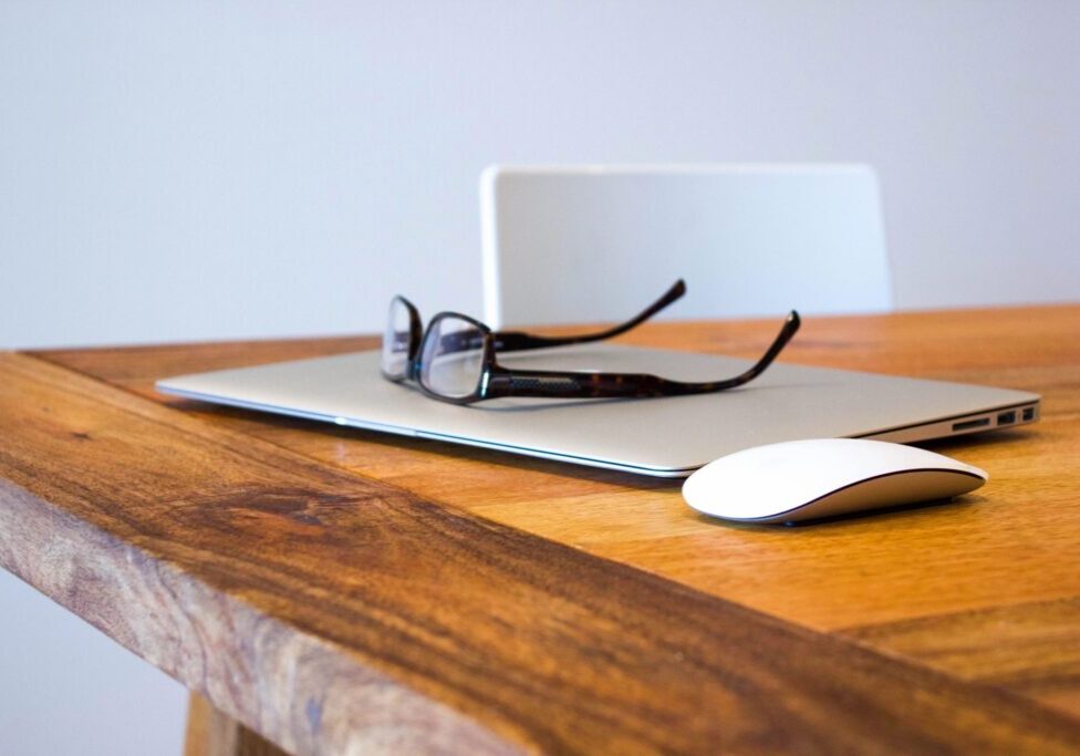 Glasses resting on a closed laptop on a wooden table with a mouse beside it.